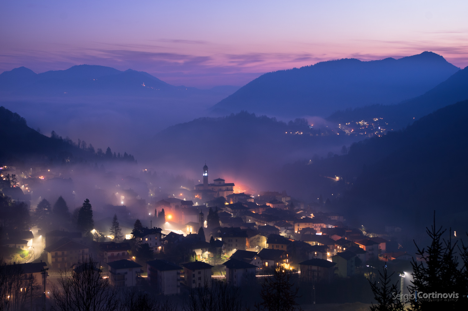 Serina (Valle Brembana - Bergamo), vista dalla strada che porta a Valpiana durante un tramonto invernale, con un sottile strato di nebbia che copre il paese