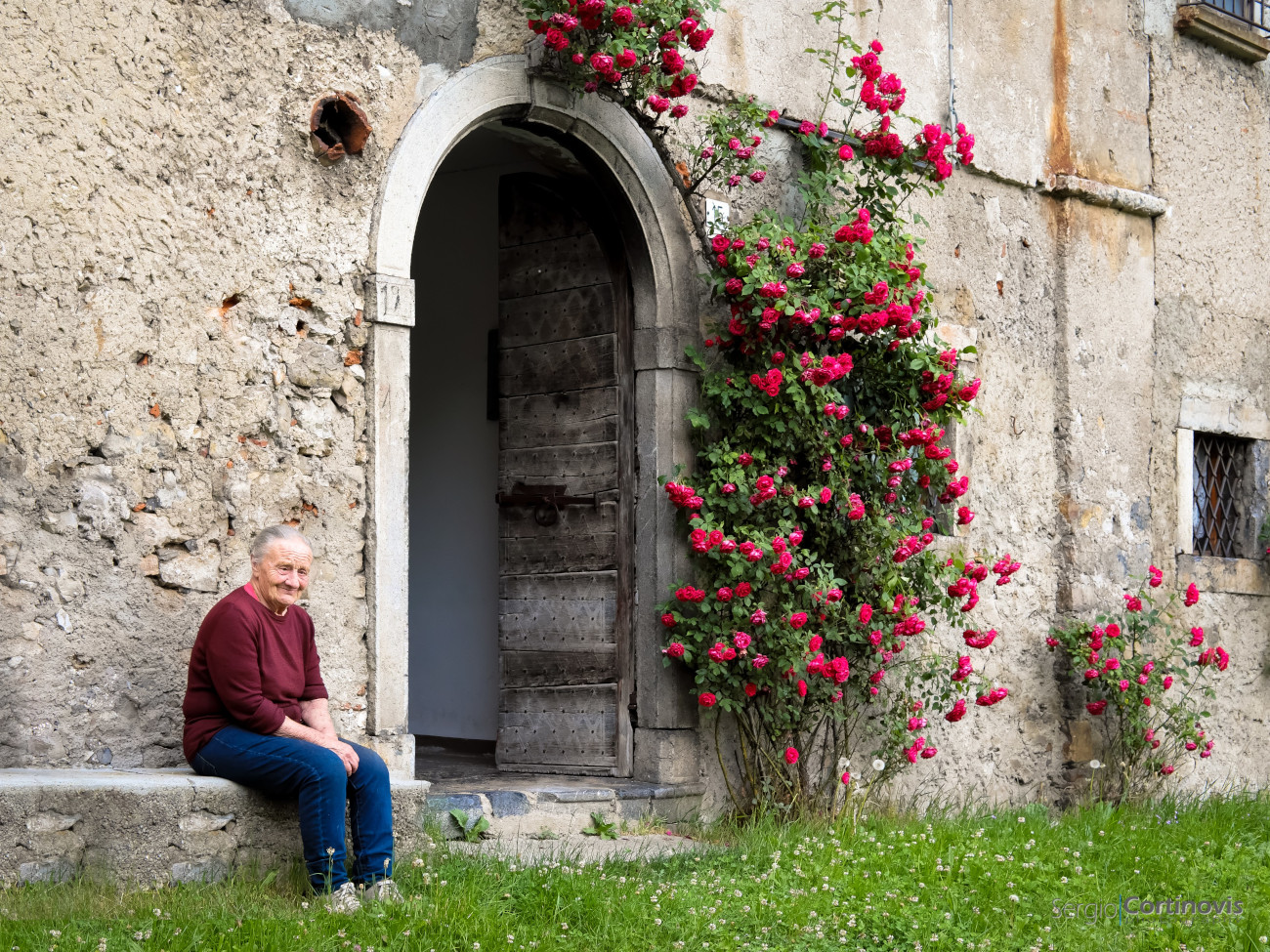 Signora anziana su una panchina con un portone e delle rose rosse sullo sfondo