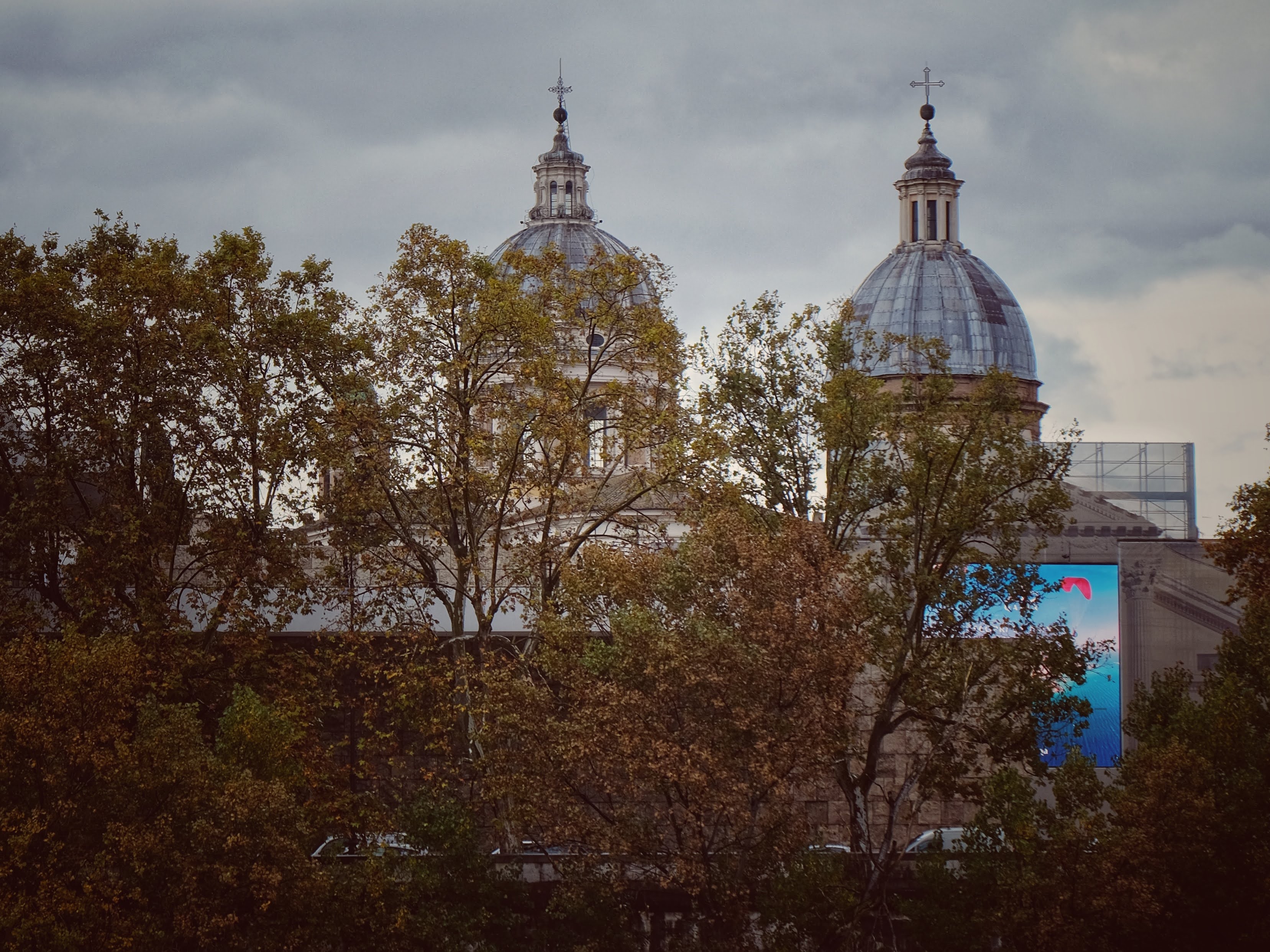 Le cupole di due Chiese di Roma dietro ad alcuni alberi dai colori autunnali, davanti ad un cielo grigio
