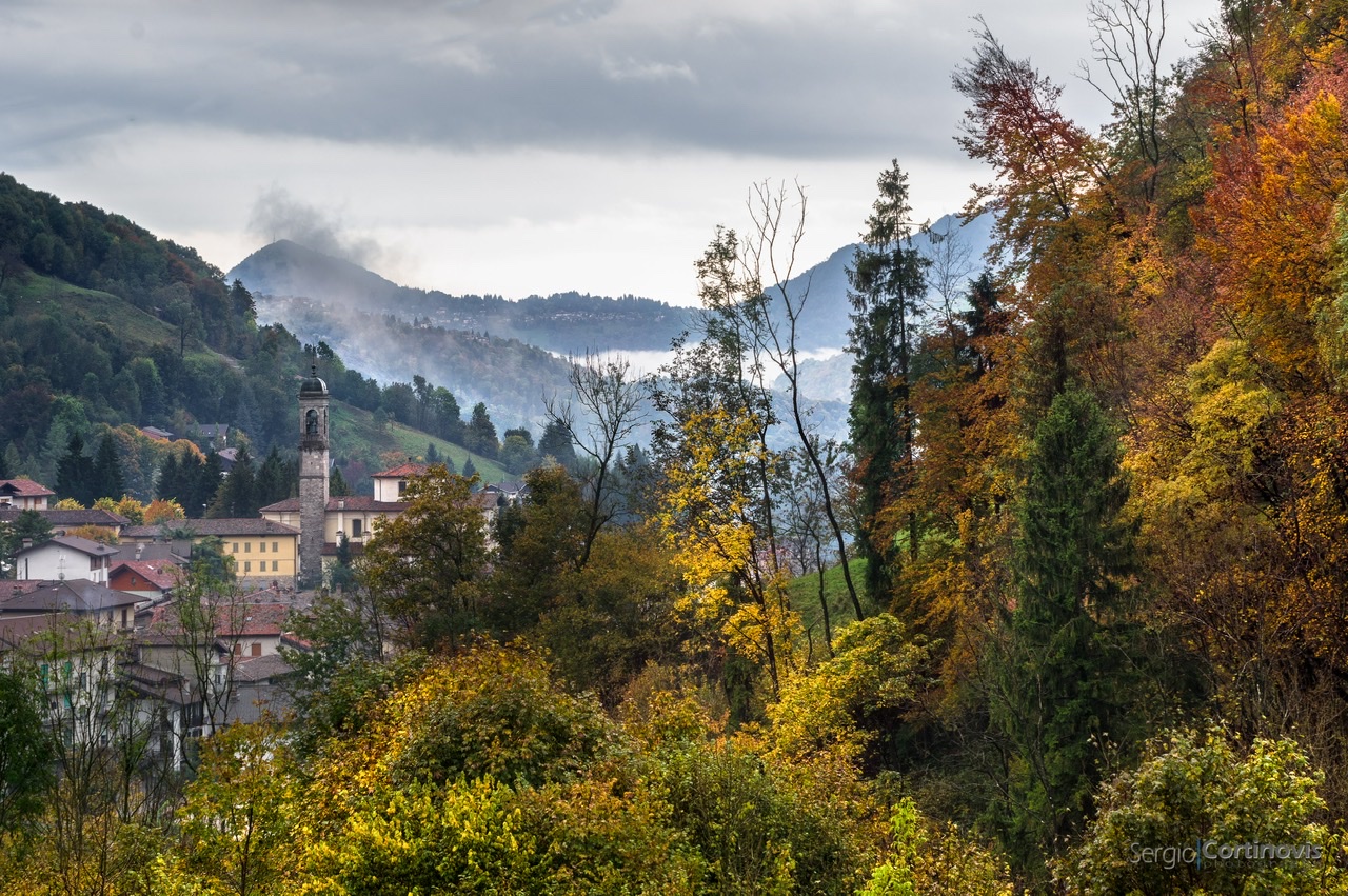 Serina in autunno, con gli alberi nel pieno dei loro colori, la pioggia e la nebbia. Sullo sfondo, la chiesa parrocchiale di Santa Maria Annunciata